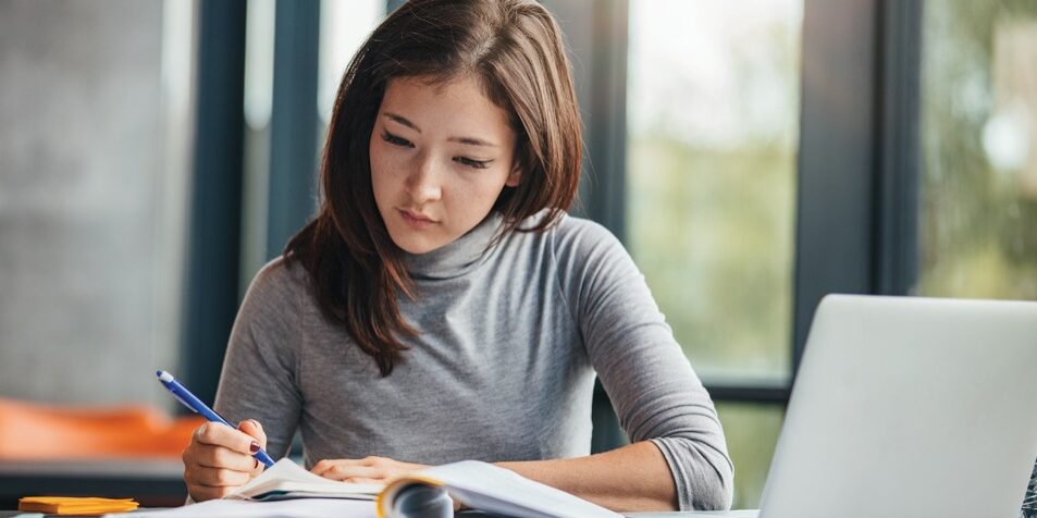 Shot of young woman taking down notes in diary. Female university student preparing note for the exam at library.